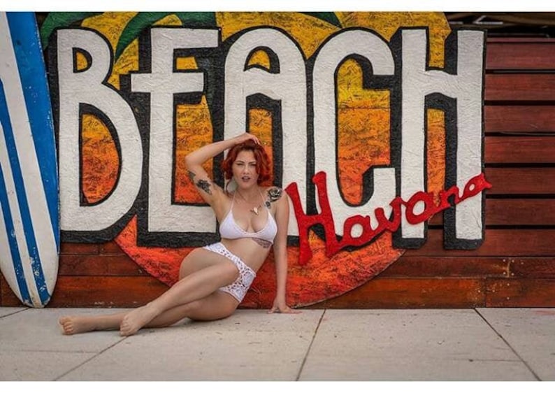 K&ouml;nnte beinhalten: Eine Frau in einem wei&szlig;en Bikini-Oberteil und wei&szlig;en H&auml;kel-H&ouml;schen posiert vor einem farbenfrohen Wandbild mit der Aufschrift "Beach Havana".
