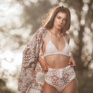 K&ouml;nnte beinhalten: Eine Frau tr&auml;gt ein wei&szlig;es Bikini-Oberteil aus H&auml;kelarbeit und ein passendes, hoch tailliertes H&auml;kel-H&ouml;schen. Sie tr&auml;gt einen floralen Kimono &uuml;ber dem Bikini. Die Frau steht in einem Wald mit verschwommenem Hintergrund.