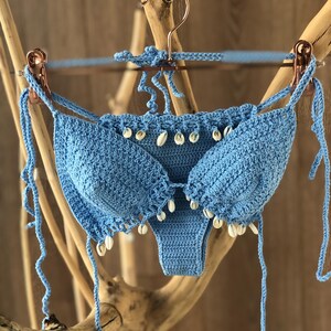 May include: A light blue crochet bikini top and bottom with white shell accents. The bikini is hanging on a wooden branch.
