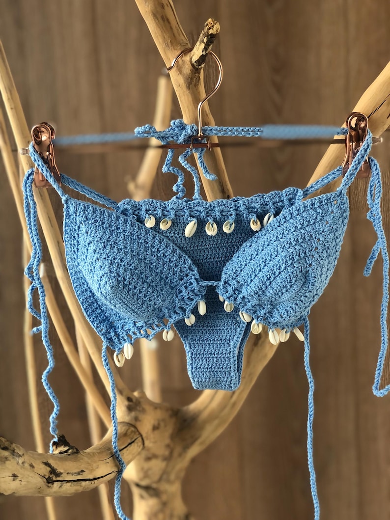 May include: A light blue crochet bikini top and bottom with white shell accents. The bikini is hanging on a wooden branch.
