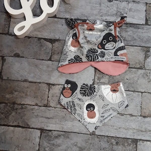 May include: A baby hat and bib set with a monkey and leaf pattern. The hat has pink ear flaps and a drawstring. The bib is triangular. The background is a brick wall with a white "Love" sign.