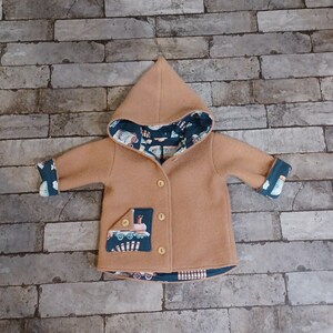 Könnte beinhalten: Ein beigefarbener Baby-Mantel mit Kapuze und Holzknöpfen. Der Mantel hat ein dunkelblaues Futter mit einem Fahrzeugmuster und einen Zugaufnäher auf der Tasche. Die Manschetten sind ebenfalls mit dem gleichen gemusterten Stoff gefüttert.