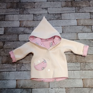Könnte beinhalten: Cremefarbener Baby-Mantel mit Kapuze und rosa gemustertem Futter. Der Mantel hat eine runde Tasche mit Mäuseaufdruck und weißen Knöpfen. Die Manschetten und das Kapuzenfutter sind rosa mit einem Mäusemuster.