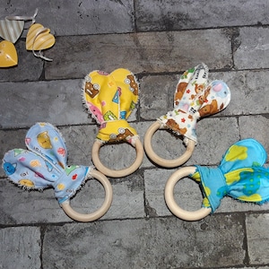 May include: Four baby teething rings with wooden rings and fabric bunny ears. The fabric features colorful patterns of cars, bears, and cats. The rings are on a gray brick surface, with heart-shaped tags in the background.