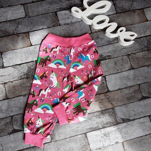 Pode incluir: Calças de pijama rosa com um padrão colorido de unicórnios e arco-íris. As calças têm cós e punhos rosa. Uma placa branca com a palavra "Love" está ao fundo.