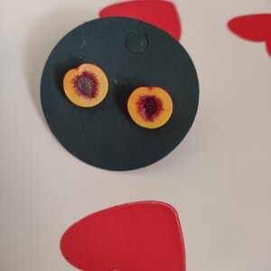 Pode incluir: Brincos de botão em forma de pêssego. Cada brinco é uma fatia de pêssego em miniatura, com um exterior amarelo e um centro vermelho-roxo, fixado em um suporte circular verde escuro. Os brincos são exibidos em uma superfície branca com formas de coração vermelhas.