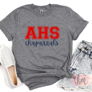 May include: A heather gray t-shirt with the red letters "AHS" and the word "chaparral" in navy blue. The shirt is paired with denim shorts and a white shoe.