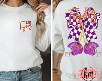 May include: White long-sleeve sweatshirt featuring the text "LSU Tigers" in gold and purple. The back of the shirt displays a purple and white checkered bow with the words "LSU Tigers" in gold. Two purple football helmets are below the bow.