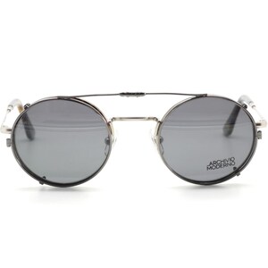 Puede incluir: Gafas de sol redondas con lentes grises y montura plateada. Las gafas tienen un doble puente y el texto "ARCHIVO MODERNO" est&aacute; impreso en la lente derecha.