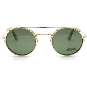 May include: Gold-tone round sunglasses with green lenses. The sunglasses have a double bridge and side accents. The text "ARCHIVO MODERNO" is printed on the right lens.