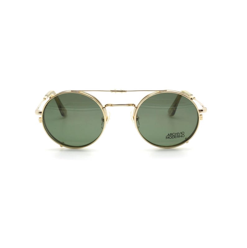 May include: Gold-tone round sunglasses with green lenses. The sunglasses have a double bridge and side accents. The text "ARCHIVO MODERNO" is printed on the right lens.