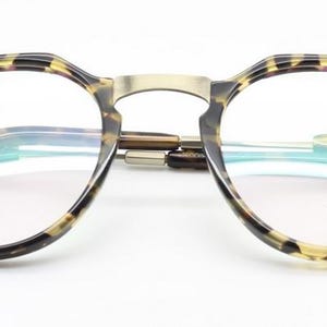 Puede incluir: Gafas redondas con montura con estampado de carey y puente dorado. Las lentes son transparentes y las patillas tienen un dise&ntilde;o sutil. El texto "ARCHIVO MODERNO" es visible en la lente.