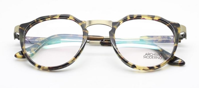 Puede incluir: Gafas redondas con montura con estampado de carey y puente dorado. Las lentes son transparentes y las patillas tienen un dise&ntilde;o sutil. El texto "ARCHIVO MODERNO" es visible en la lente.