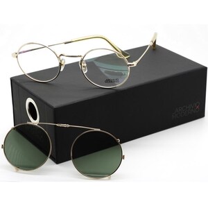 May include: Gold-rimmed eyeglasses and clip-on sunglasses rest on a black box. The eyeglasses have clear lenses and a round frame. The clip-on sunglasses have green lenses and a matching gold frame. The box has the text "ARCHIVO MODERNO".