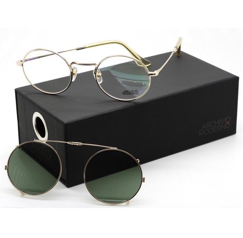 May include: Gold-rimmed eyeglasses and clip-on sunglasses rest on a black box. The eyeglasses have clear lenses and a round frame. The clip-on sunglasses have green lenses and a matching gold frame. The box has the text "ARCHIVO MODERNO".