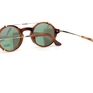 May include: Round brown sunglasses with green lenses. The glasses have a gold-colored bridge and arms, with a tortoiseshell pattern on the earpieces. The lenses have a slight green tint, and the frame is a warm brown color.