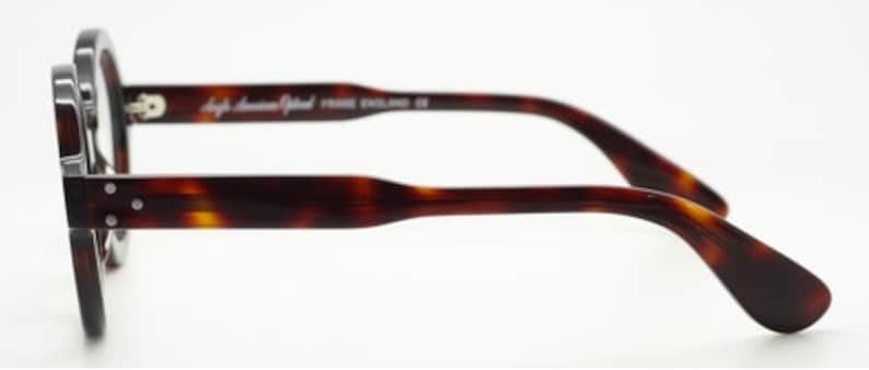 Puede incluir: Un par de gafas de pasta de carey con montura de metal plateado. Las gafas tienen forma redonda y las patillas son de pl&aacute;stico de carey. El nombre de la marca "High American Optical" est&aacute; impreso en la patilla.