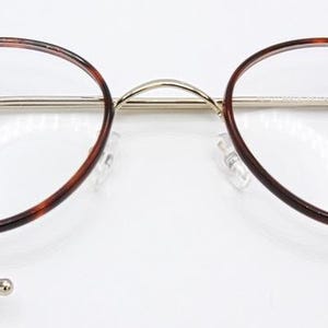 May include: Round eyeglasses with a tortoiseshell pattern on the rims and temples. The frames are a combination of gold and silver metal. The lenses are clear, and the nose pads are transparent. The glasses are set against a white background.