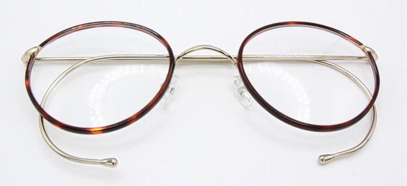 May include: Round eyeglasses with a tortoiseshell pattern on the rims and temples. The frames are a combination of gold and silver metal. The lenses are clear, and the nose pads are transparent. The glasses are set against a white background.