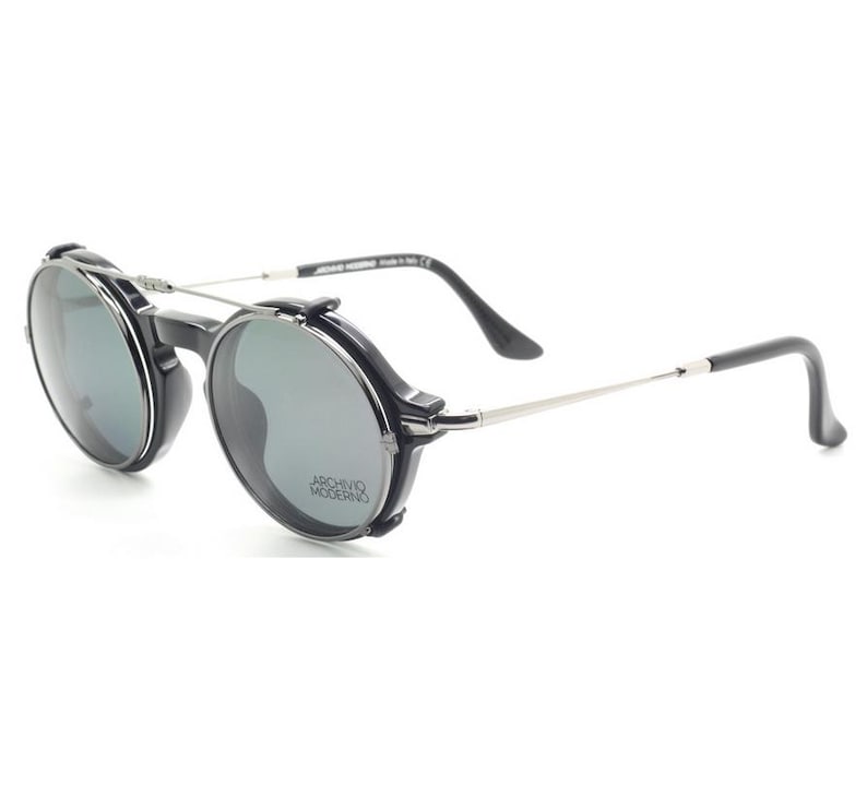 May include: Black and silver-toned sunglasses with round lenses. The sunglasses have a unique design with a black frame and silver-toned arms. The lenses are a dark gray color. The text "ARCHIVO MODERNO" is visible on the lens.