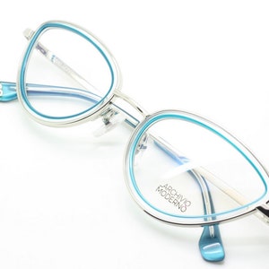 May include: Silver cat-eye glasses with a blue rim. The glasses have a clear lens and the brand name "Archivio Moderno" is printed on the right lens.