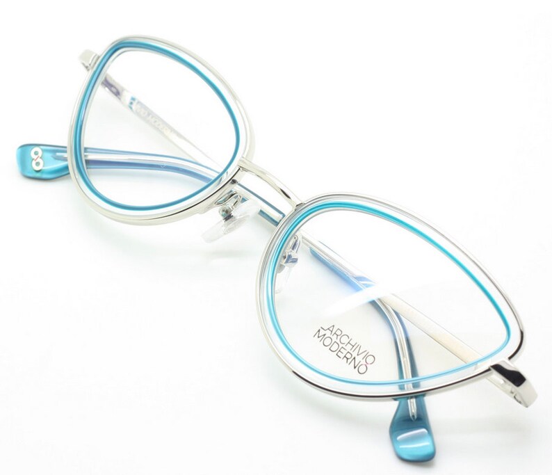 May include: Silver cat-eye glasses with a blue rim. The glasses have a clear lens and the brand name "Archivio Moderno" is printed on the right lens.