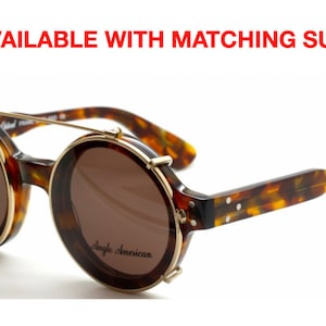 Puede incluir: Gafas de sol redondas de carey con detalles en metal dorado y un parasol de clip. Las gafas de sol tienen lentes marrones y el nombre de la marca "Angle American" est&aacute; impreso en la montura.