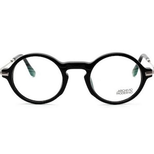May include: Black round-frame eyeglasses with silver-toned temples. The glasses have a keyhole bridge and the text "ARCHIVO MODERNO" is printed on the right lens. The lenses are clear.