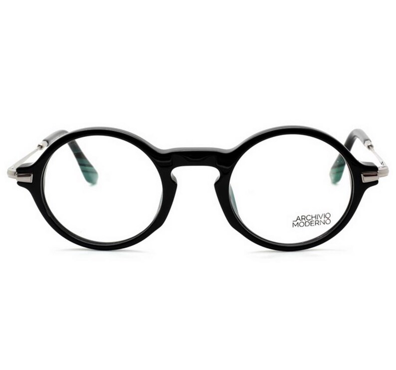 May include: Black round-frame eyeglasses with silver-toned temples. The glasses have a keyhole bridge and the text "ARCHIVO MODERNO" is printed on the right lens. The lenses are clear.