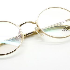 May include: Gold-rimmed eyeglasses with round lenses. The frames have a delicate design, and the temples are a light yellow color. The text "ARCHIVO MODERNO" is visible on the right temple. The glasses are set against a white background.