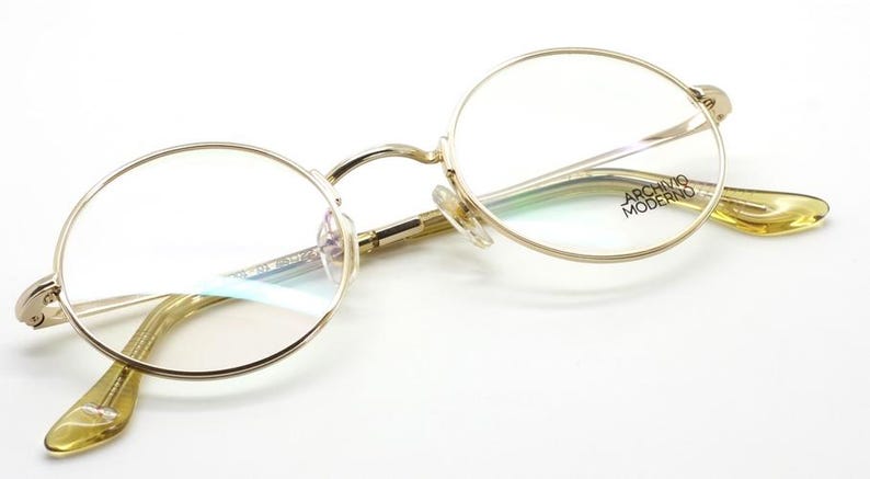 May include: Gold-rimmed eyeglasses with round lenses. The frames have a delicate design, and the temples are a light yellow color. The text "ARCHIVO MODERNO" is visible on the right temple. The glasses are set against a white background.