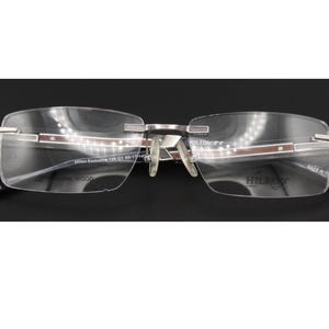 May include: Rimless eyeglasses featuring silver-tone metal temples with brown wood-grain inlays. The lenses are clear and rectangular, with the text Hilton Exclusive 129 G1 55-17-140 and Made in Italy printed on the inner surface of the lenses.