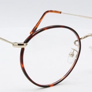 May include: Round eyeglasses with a tortoiseshell pattern on the rims and temples. The frames have a silver-colored bridge and arms. The lenses are clear. The glasses are set against a white background.