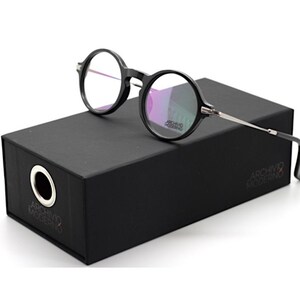 May include: Black round-frame eyeglasses with silver-toned temples rest on a black box. The eyeglasses have clear lenses. The box has the text "ARCHIVO MODERNO" printed on it. The box has a circular hole on the side.