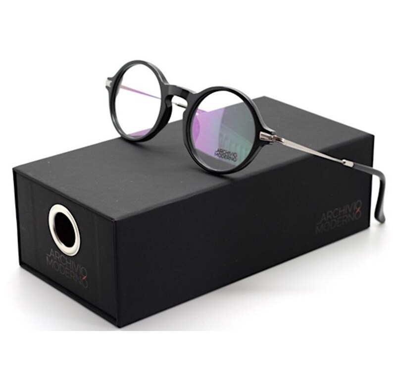 May include: Black round-frame eyeglasses with silver-toned temples rest on a black box. The eyeglasses have clear lenses. The box has the text "ARCHIVO MODERNO" printed on it. The box has a circular hole on the side.