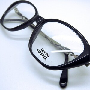 May include: Black cat-eye glasses with clear lenses and the brand name "Gianni Versace" printed on the frame.