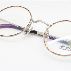 Puede incluir: Gafas redondas con montura plateada y bordes y patillas con estampado de carey. Las lentes son transparentes y las patillas tienen un estampado marr&oacute;n oscuro y beige. El texto "ARCHIVIO MODERNO" es visible en la lente.