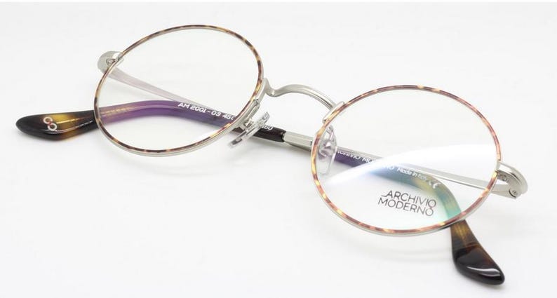 Puede incluir: Gafas redondas con montura plateada y bordes y patillas con estampado de carey. Las lentes son transparentes y las patillas tienen un estampado marr&oacute;n oscuro y beige. El texto "ARCHIVIO MODERNO" es visible en la lente.