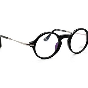 May include: Black round-frame eyeglasses with silver-toned temples. The glasses have a classic design with a glossy finish. The lenses are clear, and the frame is thick. The glasses are set against a white background.
