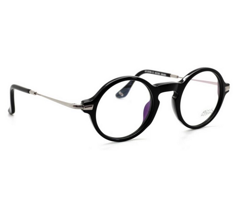 May include: Black round-frame eyeglasses with silver-toned temples. The glasses have a classic design with a glossy finish. The lenses are clear, and the frame is thick. The glasses are set against a white background.