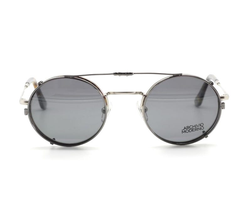 Puede incluir: Gafas de sol redondas con lentes grises y montura plateada. Las gafas tienen un doble puente y el texto "ARCHIVO MODERNO" est&aacute; impreso en la lente derecha.