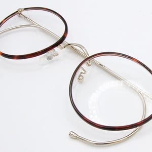 May include: Round eyeglasses with a tortoiseshell pattern on the rims and temples. The frames are a combination of gold-tone metal and clear plastic. The lenses are clear, and the glasses are open and resting on a white surface.