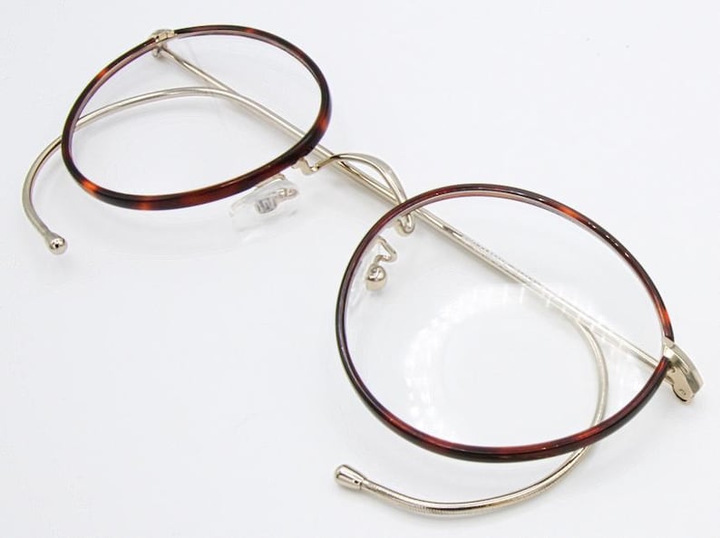 May include: Round eyeglasses with a tortoiseshell pattern on the rims and temples. The frames are a combination of gold-tone metal and clear plastic. The lenses are clear, and the glasses are open and resting on a white surface.