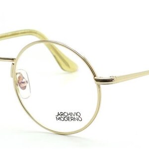 May include: Gold-tone, round-framed eyeglasses with clear lenses. The frames have a thin, delicate design with a subtle shine. The temples are a light, translucent color. The text "ARCHIVO MODERNO" is printed on the lens.