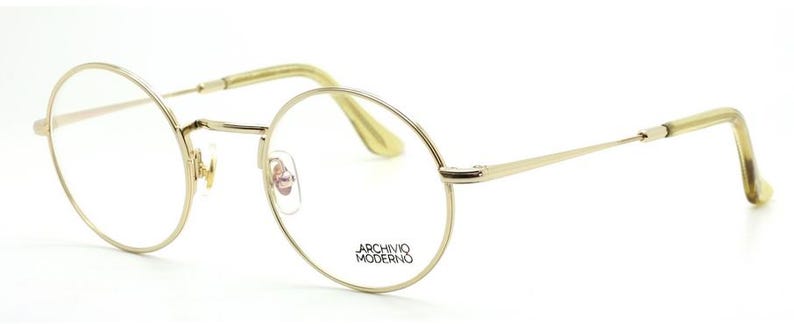 May include: Gold-tone, round-framed eyeglasses with clear lenses. The frames have a thin, delicate design with a subtle shine. The temples are a light, translucent color. The text "ARCHIVO MODERNO" is printed on the lens.