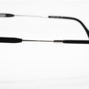 Op de afbeelding: Zwarte bril met een rechthoekig montuur en zilverkleurige veren. De bril heeft een strak, modern design met een focus op minimalistische esthetiek. De veren buigen zachtjes naar de oren.