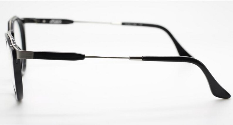Op de afbeelding: Zwarte bril met een rechthoekig montuur en zilverkleurige veren. De bril heeft een strak, modern design met een focus op minimalistische esthetiek. De veren buigen zachtjes naar de oren.