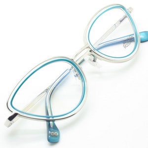 May include: Silver cat eye glasses with a blue trim. The brand name "ARCURO MODERNO" is printed on the right temple.