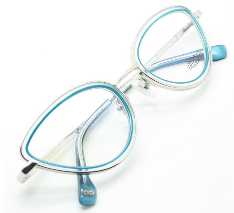 May include: Silver cat eye glasses with a blue trim. The brand name "ARCURO MODERNO" is printed on the right temple.