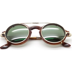 May include: Round sunglasses with a brown tortoiseshell frame and green lenses. The sunglasses have a gold-colored metal bridge and accents. The lenses have the text "ARCHIVO MUSEO" printed on them. The sunglasses are open and ready to wear.